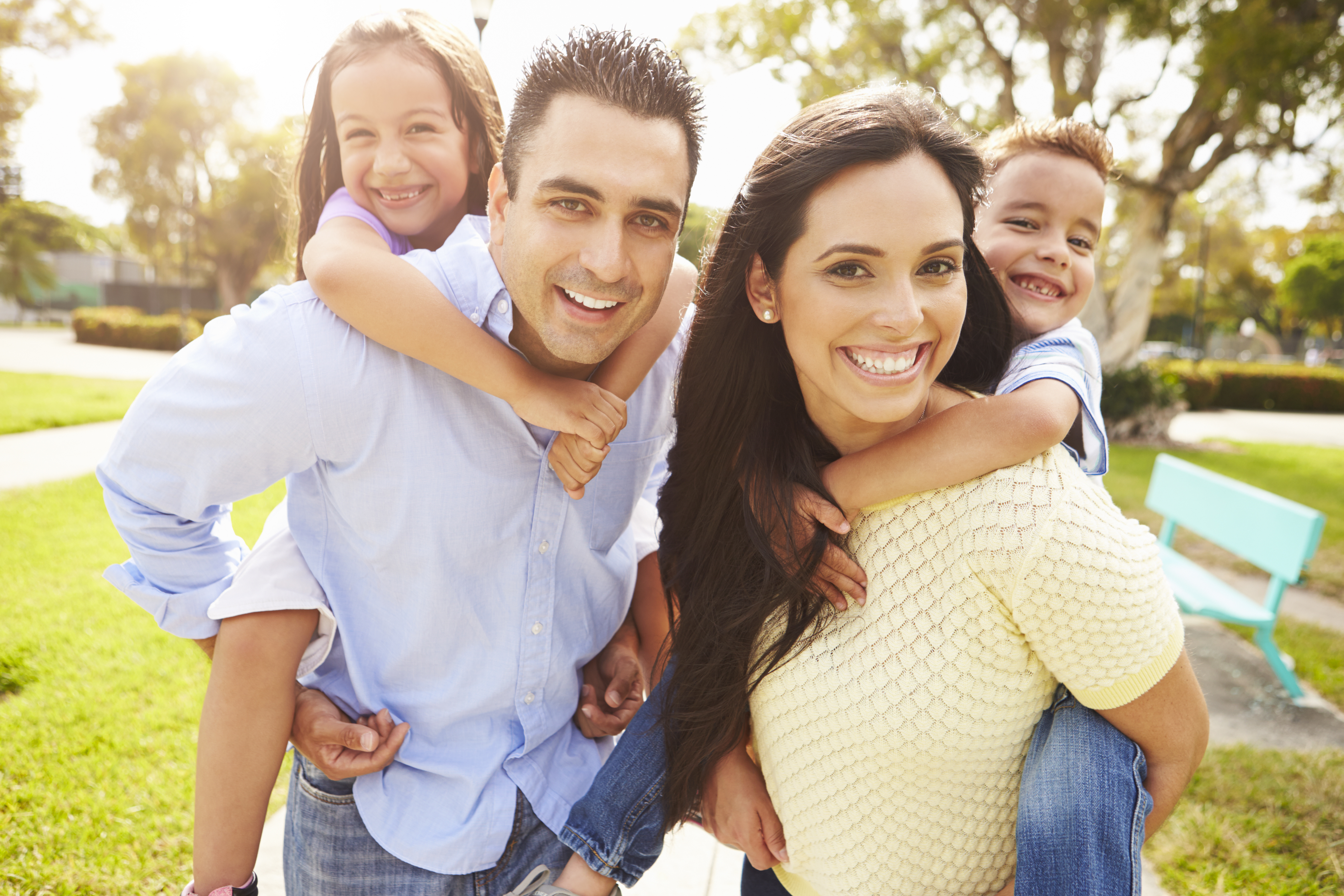 Hispanic Parents Giving Children Piggyback Ride In Garden - J Carcamo